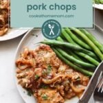 A plate with smothered pork chops topped with onions and a side of green beans sits beside a fork and knife, offering a comforting meal—perfect alongside oven baked salmon in foil for added variety.