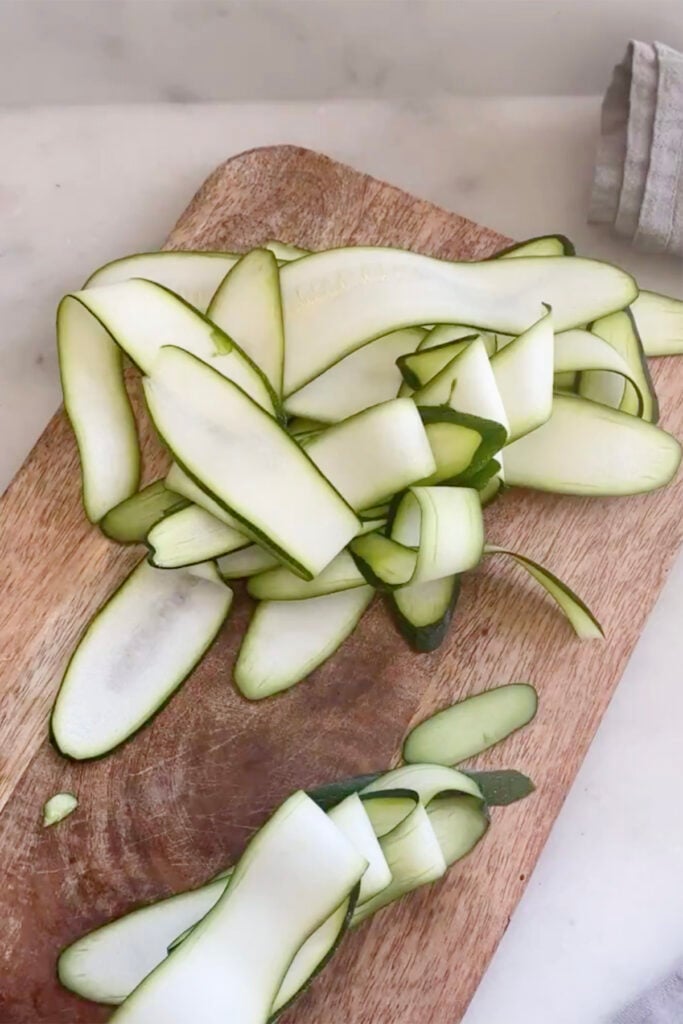 Thinly sliced zucchini strips are scattered on a wooden cutting board with a cloth visible in the background.