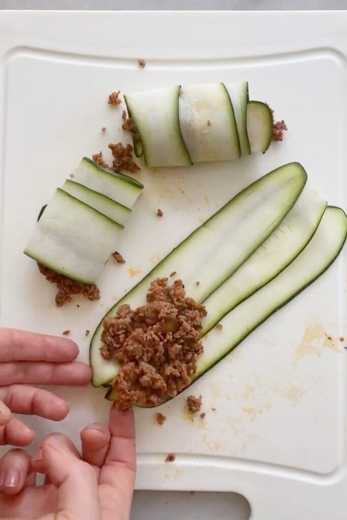 Sliced zucchini strips being filled with ground meat and rolled up on a white cutting board.