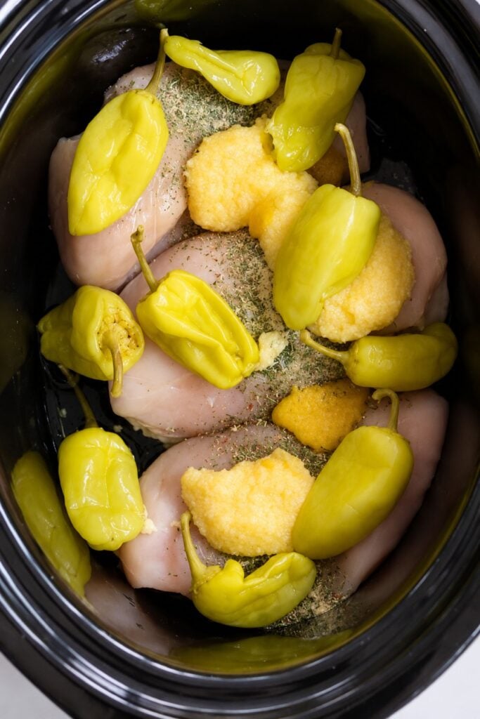 Raw chicken breasts in a slow cooker topped with pepperoncini peppers, dry seasoning, and a scoop of what appears to be butter or margarine—classic ingredients for making Mississippi chicken.