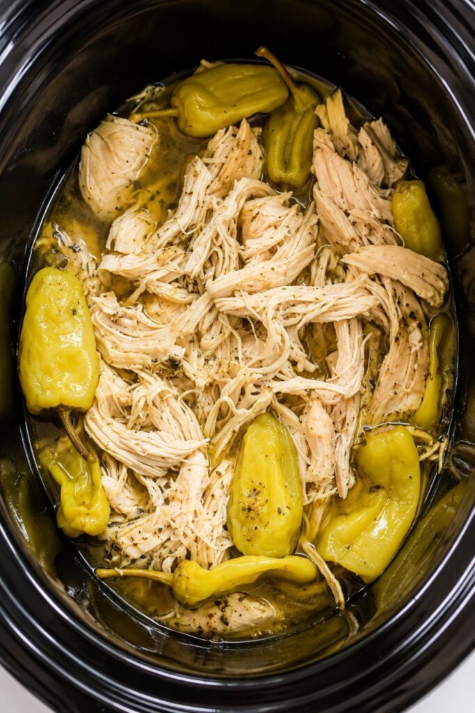 Mississippi chicken—shredded chicken and whole pepperoncini peppers simmering in a savory broth inside a black slow cooker.