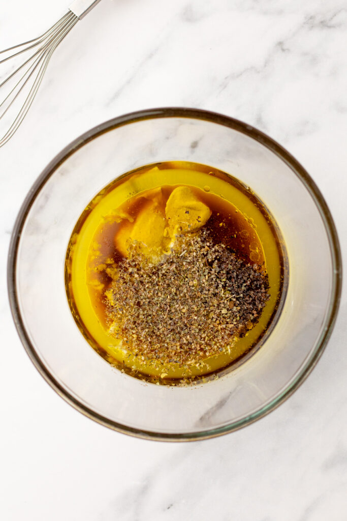 A glass bowl containing olive oil, mustard, black pepper, and seasonings sits on a white marble surface next to a metal whisk.