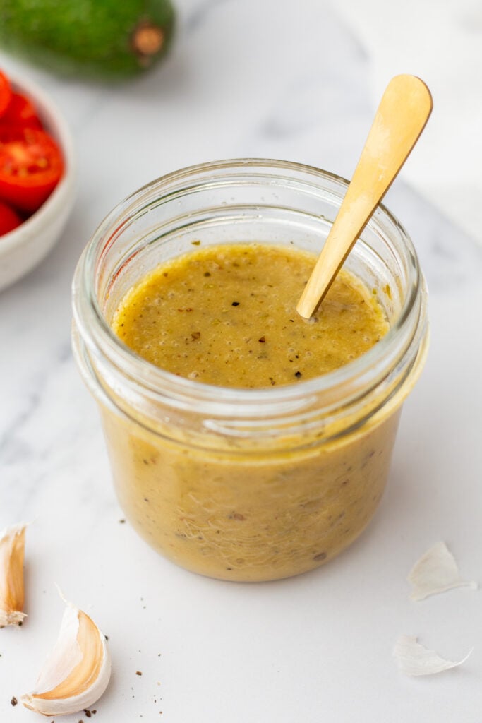 A glass jar filled with green salsa or sauce, with a gold spoon inside. Garlic cloves and a bowl of cherry tomatoes are nearby on a white surface.