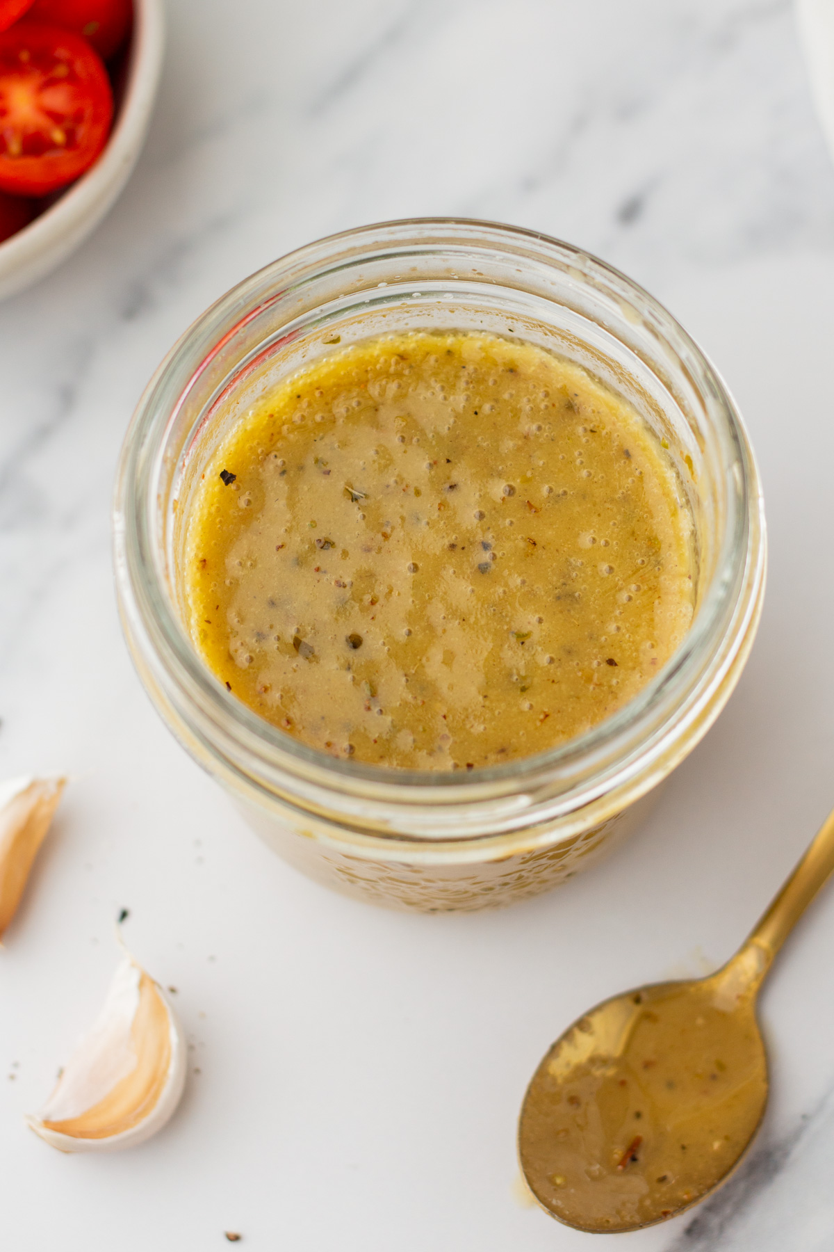 An open glass jar filled with vinaigrette dressing sits on a white surface beside a spoon, garlic cloves, and a bowl of tomatoes.