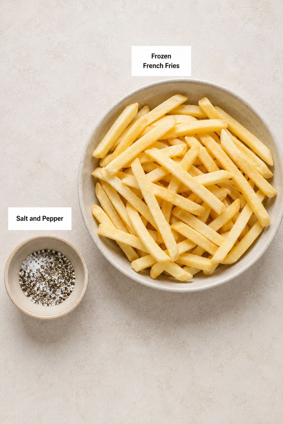 A bowl of Air Fryer Frozen French Fries sits next to a small dish of salt and pepper, both labeled on a light surface.