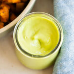 A glass jar filled with creamy green, avocado dressing sits on a light surface next to a bowl of roasted sweet potatoes and a blue cloth napkin.