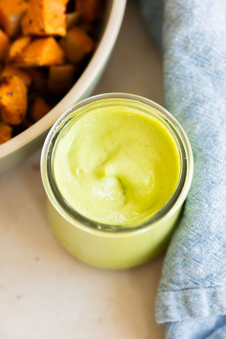 A glass jar filled with creamy green, avocado dressing sits on a light surface next to a bowl of roasted sweet potatoes and a blue cloth napkin.