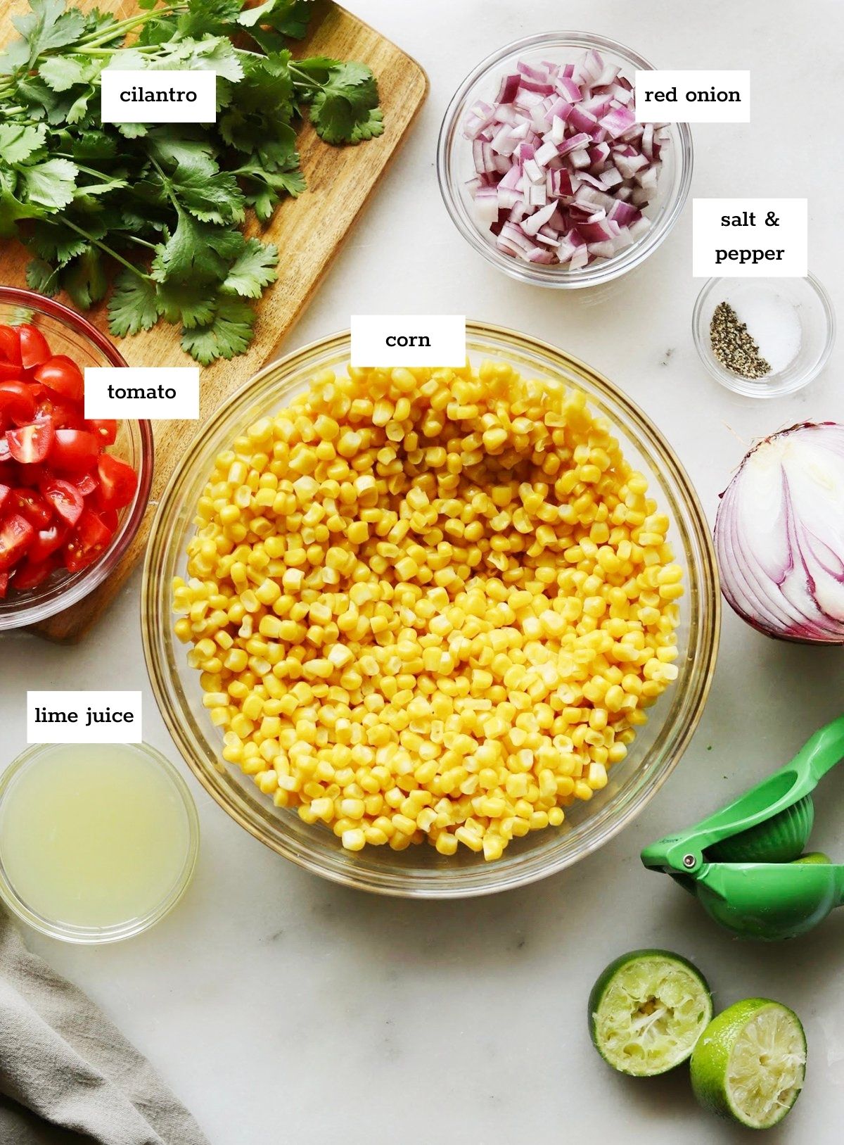 A bowl of Chipotle Corn Salsa surrounded by chopped red onion, tomato, cilantro, lime juice, salt, pepper, and a halved lime on a white surface with labels for each ingredient—just like the chipotle copycat corn salsa recipe.