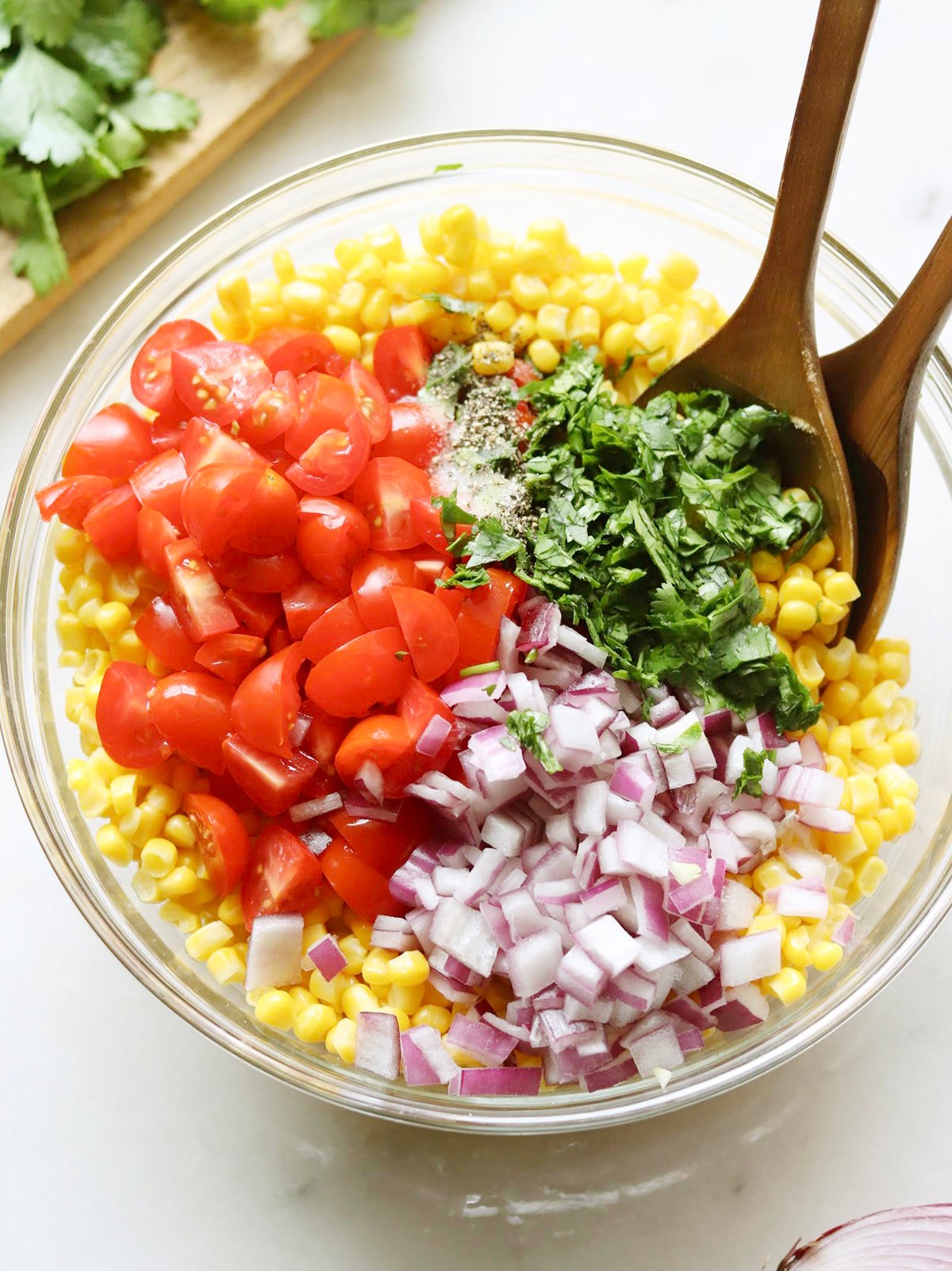 A glass bowl filled with corn, chopped tomatoes, diced red onion, and chopped cilantro, with wooden spoons placed inside.