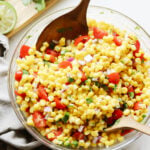 A glass bowl filled with corn salad containing corn kernels, chopped tomatoes, red onions, cilantro, and two wooden serving spoons.