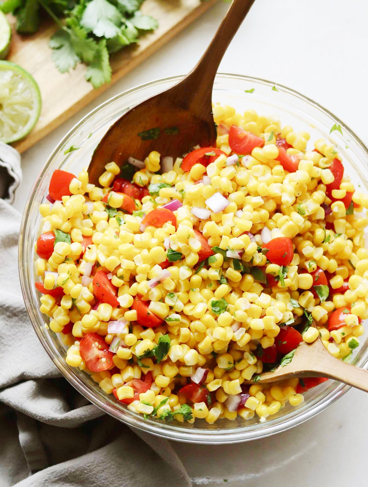 A glass bowl filled with corn salad containing corn kernels, chopped tomatoes, red onions, cilantro, and two wooden serving spoons.