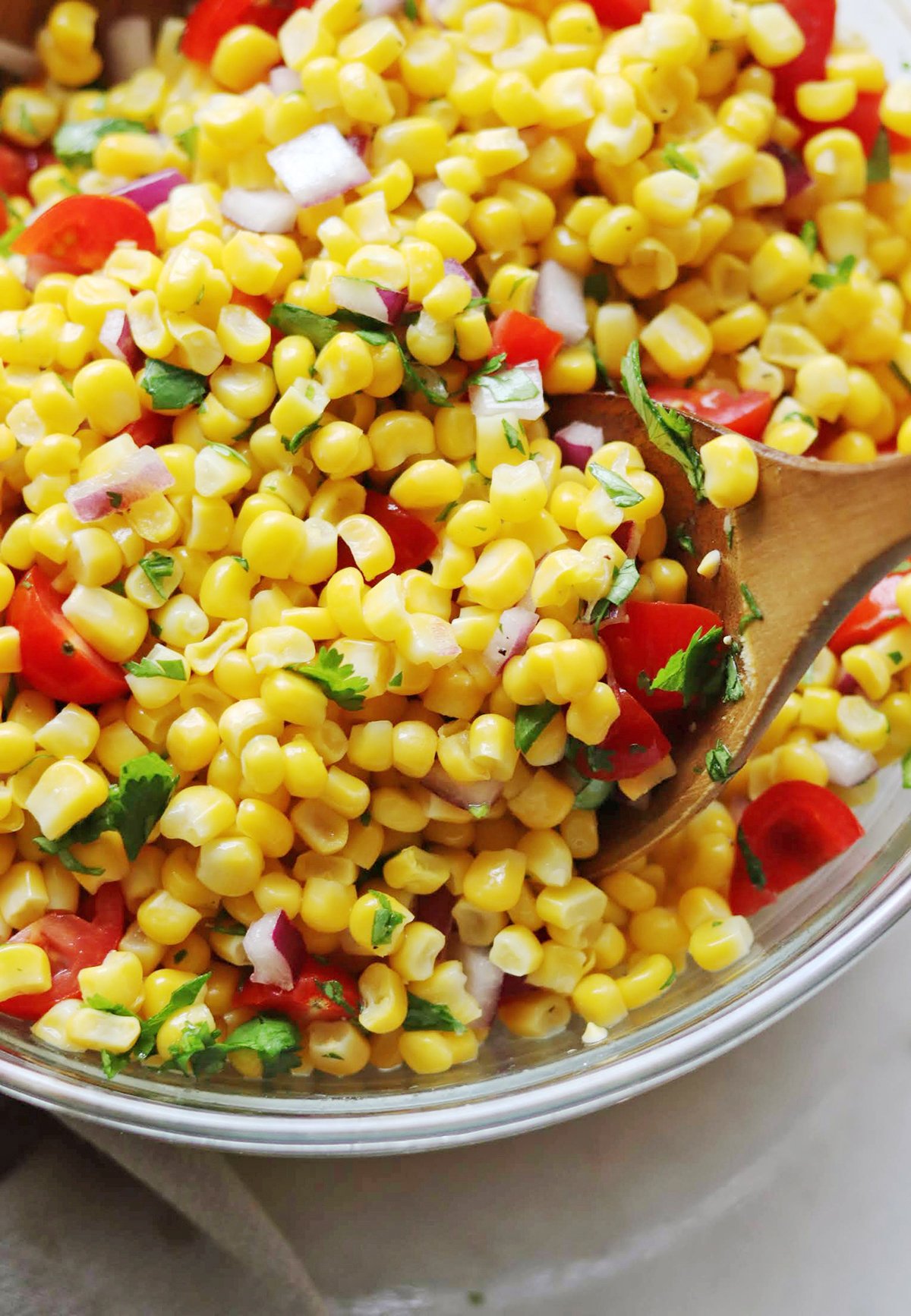 A bowl of corn salad with diced red onion, cherry tomatoes, and chopped cilantro, mixed with a wooden spoon.