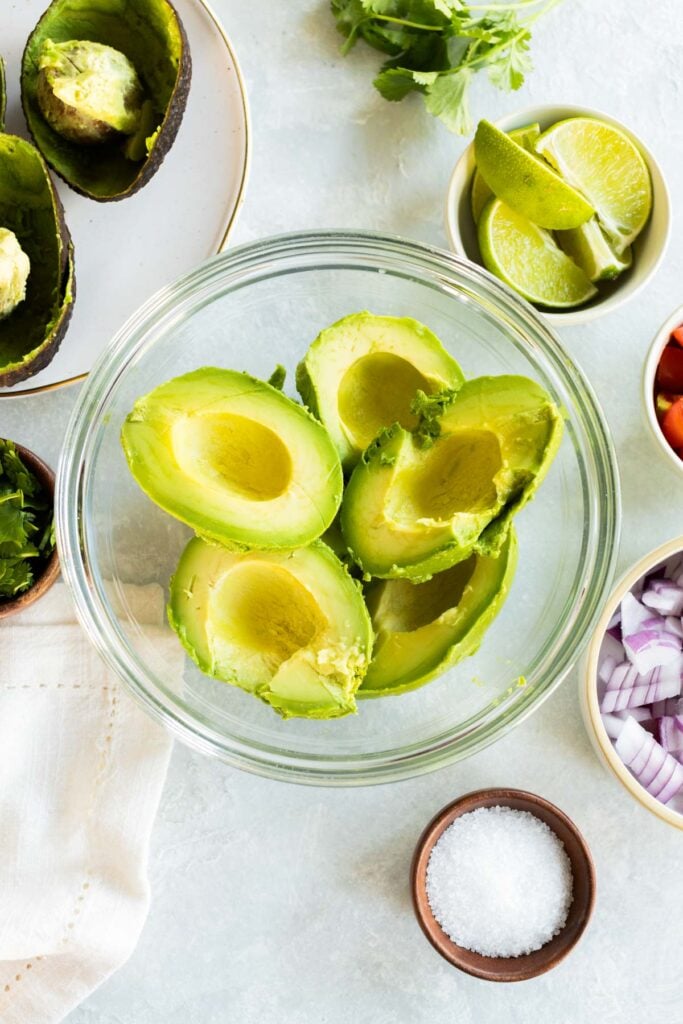 A glass bowl with halved avocados surrounded by bowls of salt, chopped red onion, lime wedges, chopped cilantro, and diced tomatoes on a white countertop.