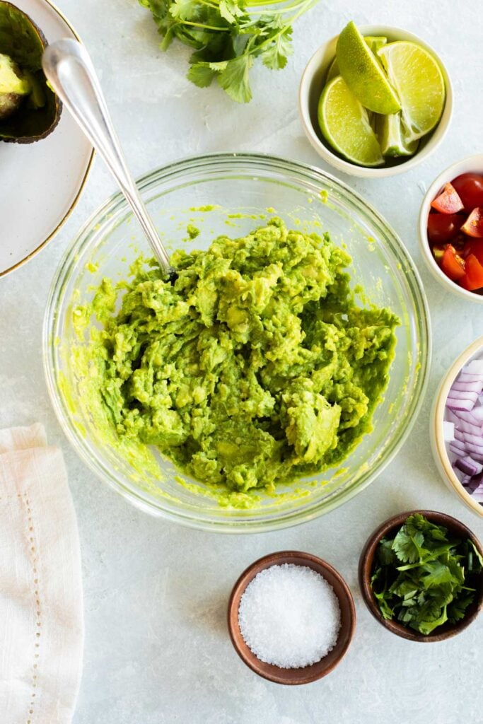 A bowl of mashed guacamole with a spoon, surrounded by bowls of lime wedges, chopped tomatoes, red onion, cilantro, and salt on a light surface.