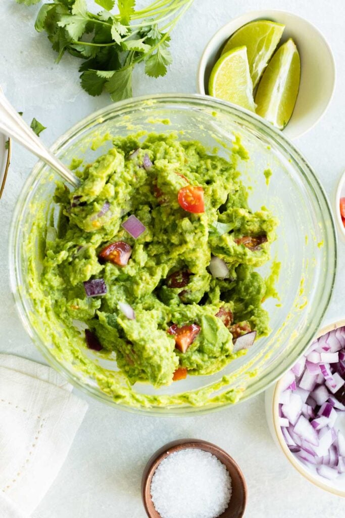 A bowl of guacamole with chopped tomatoes and red onions, surrounded by cilantro, lime wedges, salt, and diced red onions.