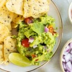 A plate of tortilla chips with guacamole garnished with cilantro, diced red onion, tomatoes, and a lime wedge, surrounded by bowls of lime, salsa, and chopped onions.