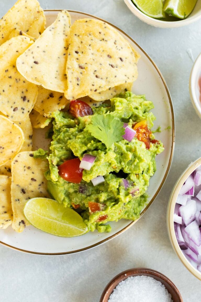 A plate of tortilla chips with guacamole garnished with cilantro, diced red onion, tomatoes, and a lime wedge, surrounded by bowls of lime, salsa, and chopped onions.