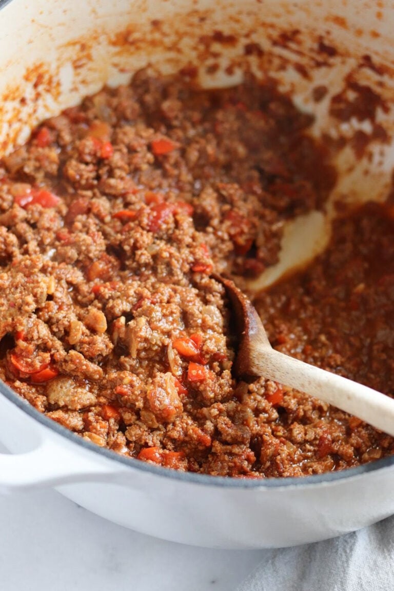 Ground beef and diced tomatoes simmer in a white pot, being stirred with a wooden spoon.
