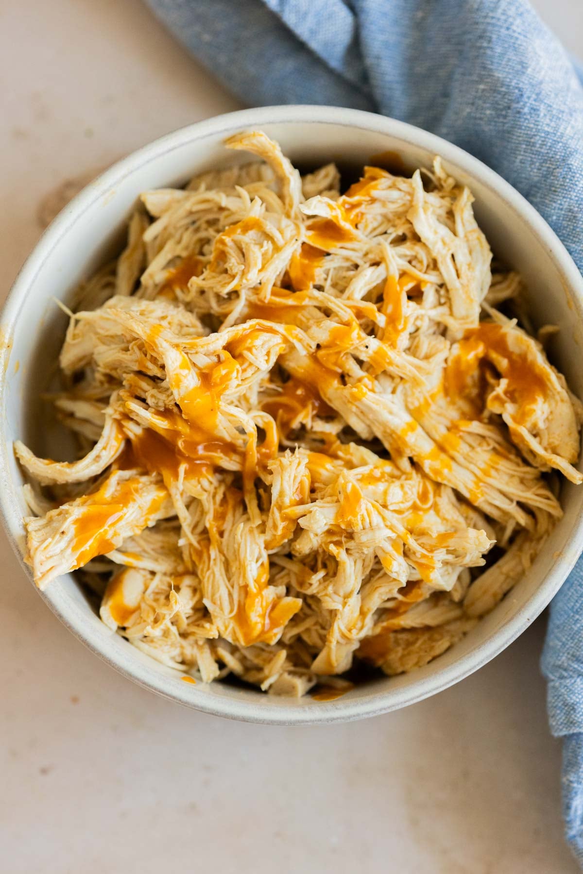 A bowl of shredded chicken with a drizzle of buffalo sauce, placed next to a blue cloth on a light surface.