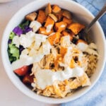 A bowl with buffalo chicken, brown rice, roasted sweet potatoes, chopped lettuce, tomatoes, and a creamy white sauce, with a spoon and a blue cloth nearby.