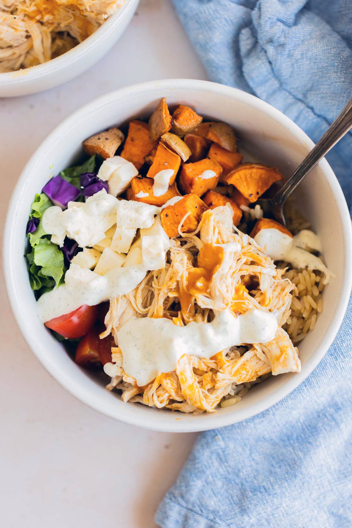 A bowl with buffalo chicken, brown rice, roasted sweet potatoes, chopped lettuce, tomatoes, and a creamy white sauce, with a spoon and a blue cloth nearby.
