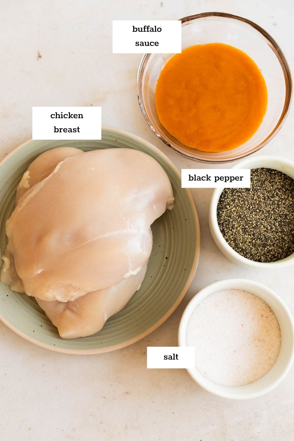 A plate with raw chicken breasts, a bowl of buffalo sauce, black pepper, and salt are arranged on a countertop—perfect ingredients for making flavorful instant pot buffalo chicken.