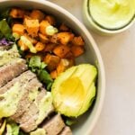 A bowl with sliced steak, avocado, roasted sweet potatoes, leafy greens, and green dressing, next to a jar of the same dressing.