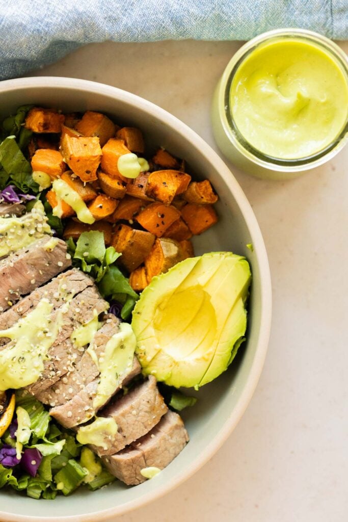A bowl with sliced steak, avocado, roasted sweet potatoes, leafy greens, and green dressing, next to a jar of the same dressing.