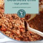 A pot of hearty beanless chili with ground meat and red bell peppers, stirred with a wooden spoon. Text overlay reads "beanless chili - high protein - cookathomemom.com".