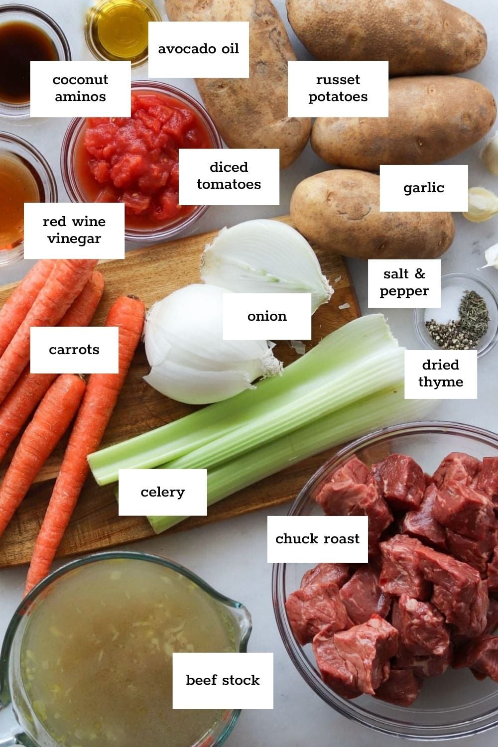 Chopped chuck roast, russet potatoes, carrots, celery, onion, garlic, diced tomatoes, beef stock, coconut aminos, red wine vinegar, avocado oil, dried thyme, salt, and pepper arranged on a counter.
