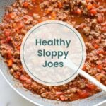 Ground beef and diced red peppers simmer in a skillet with tomato sauce for these healthy sloppy joes. A white spoon rests in the pan. Text overlay reads "Paleo Sloppy Joe.