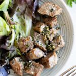A plate with air fryer steak bites topped with herbs and sea salt, served alongside mixed leafy greens. A fork and knife are placed on the side.