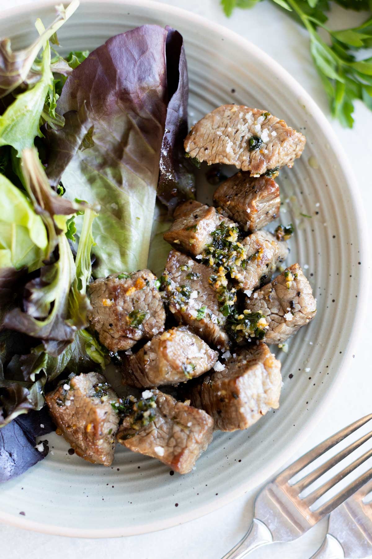 A plate with air fryer steak bites topped with herbs and sea salt, served alongside mixed leafy greens. A fork and knife are placed on the side.