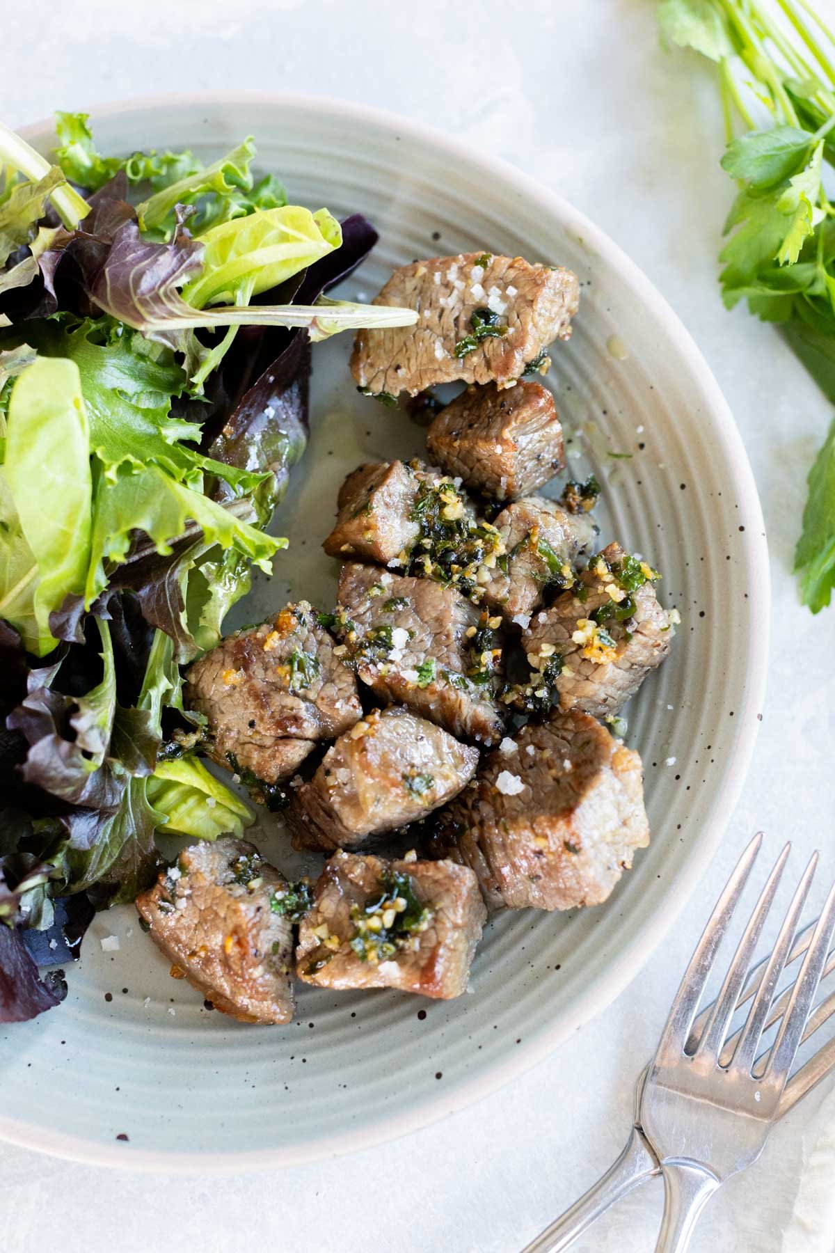 A plate with seasoned air fryer steak bites and a mixed green salad, garnished with herbs. Forks are placed beside the plate.