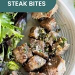 A plate with tender air fryer steak bites garnished with herbs and flaky salt, served next to mixed greens. A label reads "Air Fryer Steak Bites.