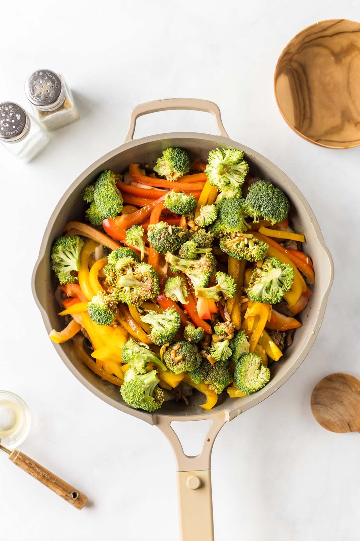A beige skillet filled with sliced red, yellow, and orange bell peppers, broccoli florets, and onions—ingredients perfect for a colorful beef stir fry—sits on a white surface, surrounded by kitchen utensils and bowls.