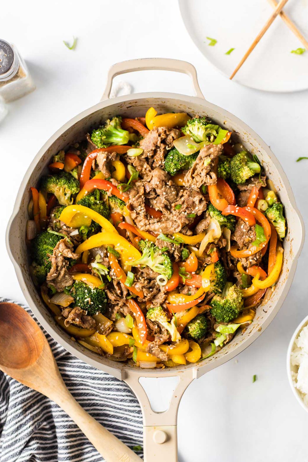 A skillet filled with savory beef stir fry, broccoli, yellow and red bell peppers, and onions, garnished with sesame seeds. A wooden spoon rests beside a plate, ready to serve this flavorful dish.