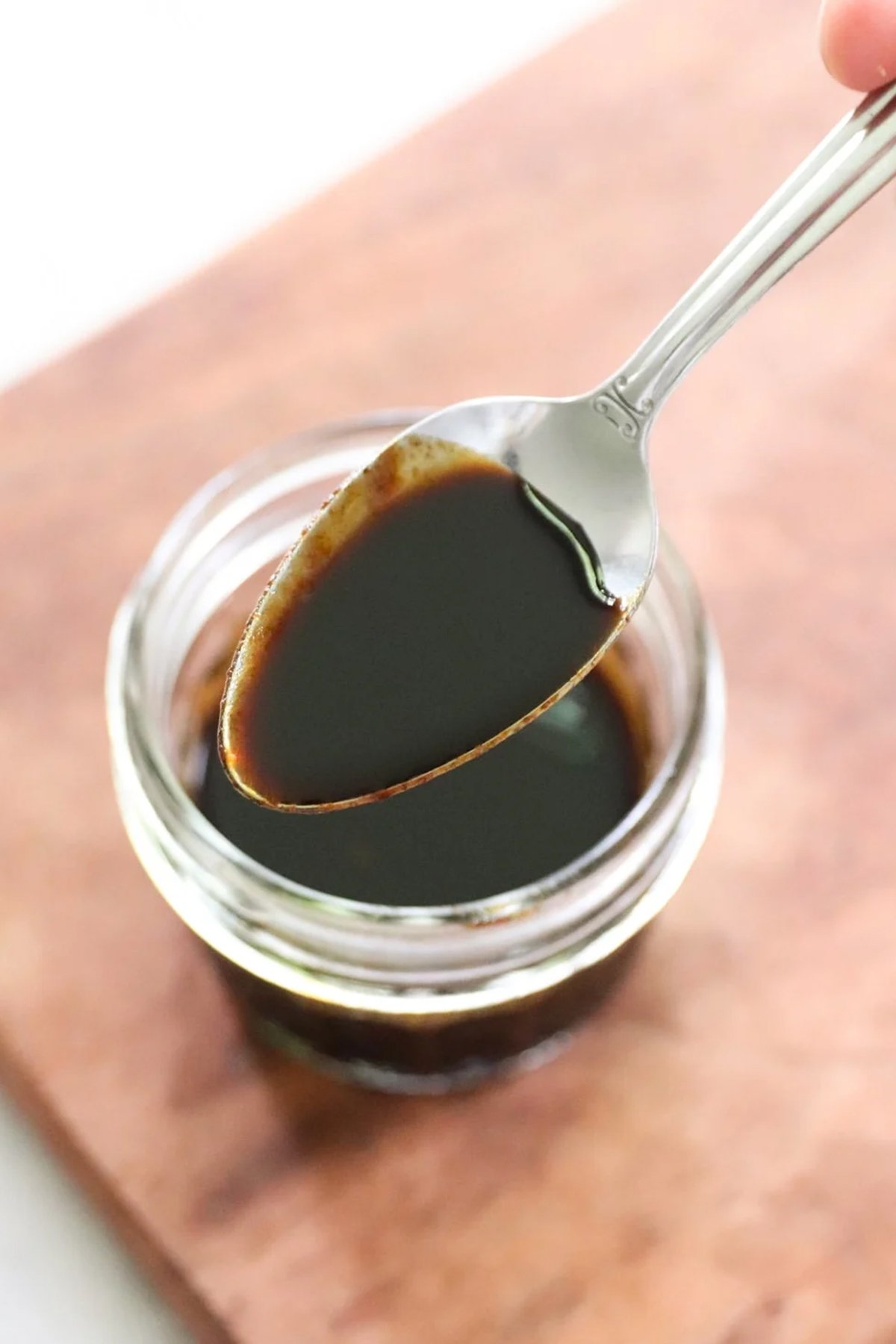 A spoon holding a small amount of dark liquid, perfect for beef stir fry, is positioned over a glass jar filled with the same liquid, set on a wooden surface.