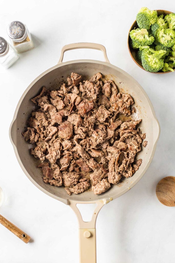 Cooked beef stir fry strips in a beige skillet on a white surface, with a bowl of broccoli florets and salt and pepper shakers nearby.