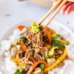 A hand using chopsticks to pick up beef, broccoli, and bell peppers from a plate of beef stir-fry.