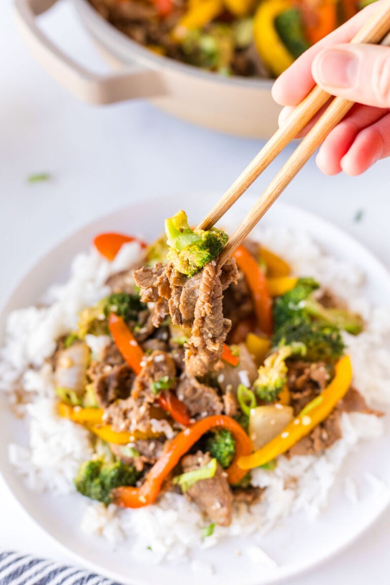 A hand using chopsticks to pick up beef, broccoli, and bell peppers from a plate of beef stir-fry.