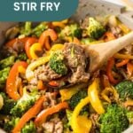 A pan of beef stir fry with broccoli, red and yellow bell peppers, and sliced beef being stirred with a wooden spoon. This colorful beef stir fry is packed with fresh veggies and savory flavors.