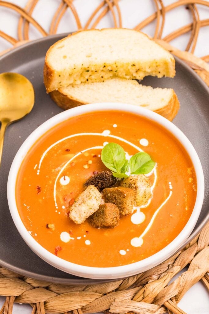 A bowl of carrot soup with cream, croutons, and basil garnish, served with two slices of bread on a gray plate. A gold spoon is placed beside the bowl.