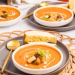A bowl of carrot soup topped with croutons and cream, served with a slice of toasted bread on a gray plate; tomatoes and another bowl in the background.