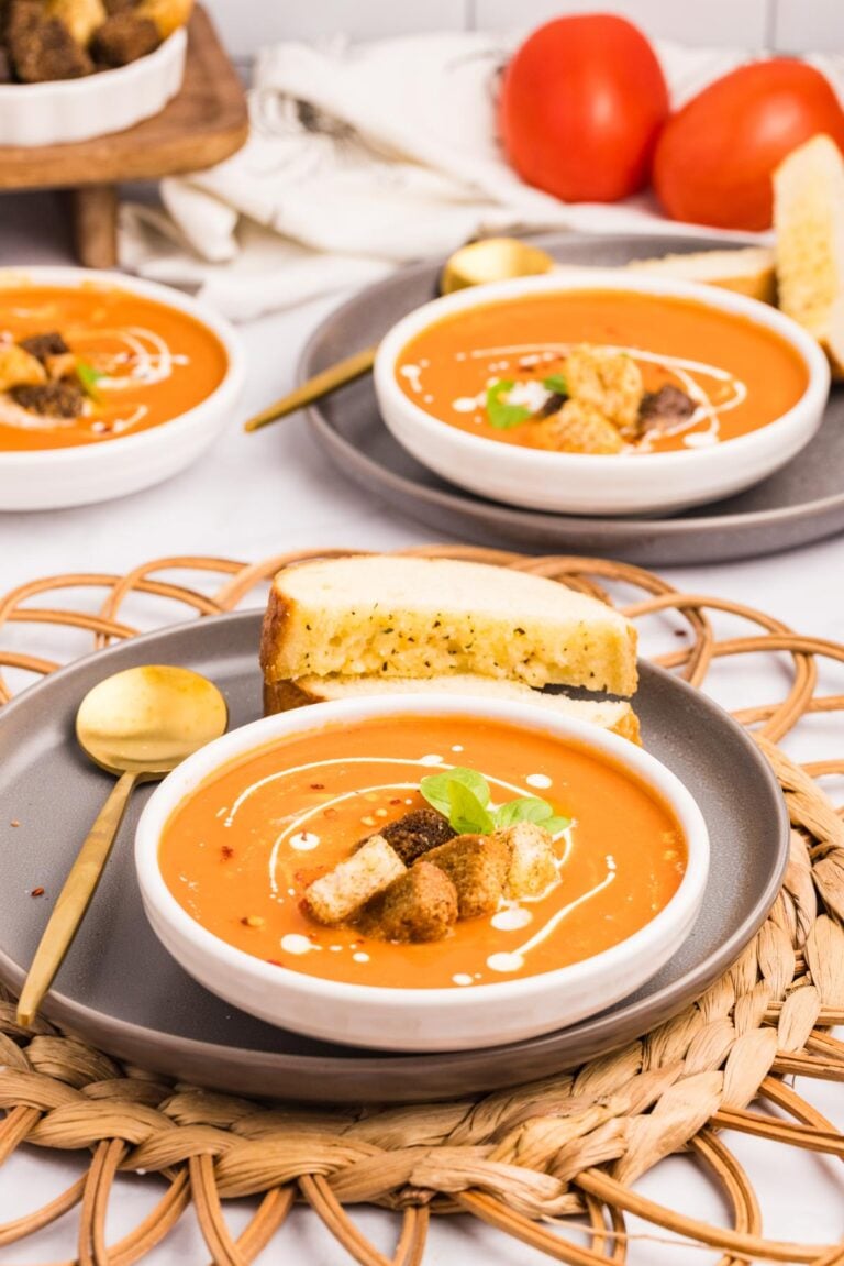 A bowl of carrot soup topped with croutons and cream, served with a slice of toasted bread on a gray plate; tomatoes and another bowl in the background.