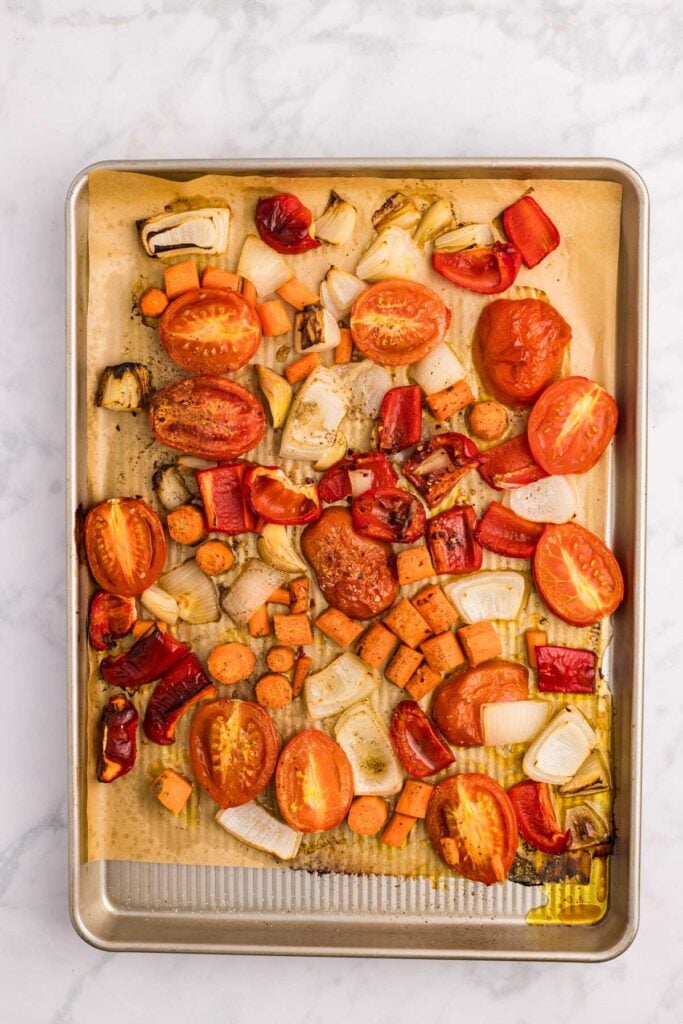 A baking sheet lined with parchment paper holds roasted vegetables, including tomatoes, onions, carrots, and red bell peppers, on a marble surface.