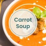A bowl of carrot soup garnished with cream, basil leaves, and croutons sits on a plate next to a gold spoon and a slice of bread, making this carrot soup the perfect comforting meal.
