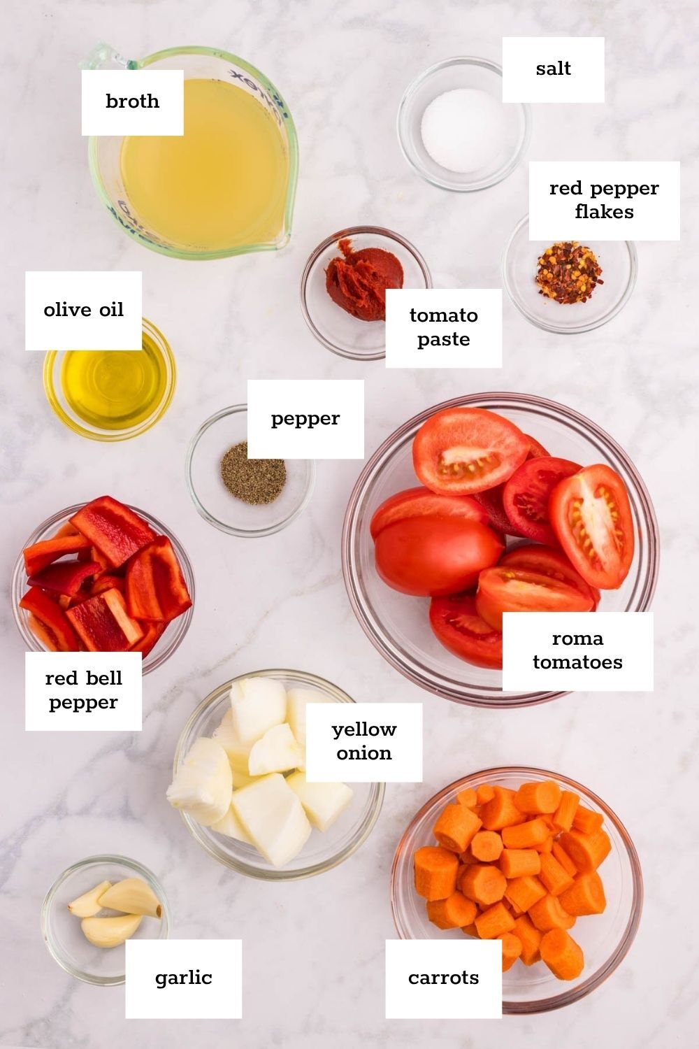 Various labeled ingredients for carrot soup are arranged on a countertop, including broth, olive oil, red bell pepper, garlic, yellow onion, carrots, roma tomatoes, tomato paste, salt, pepper, and red pepper flakes.