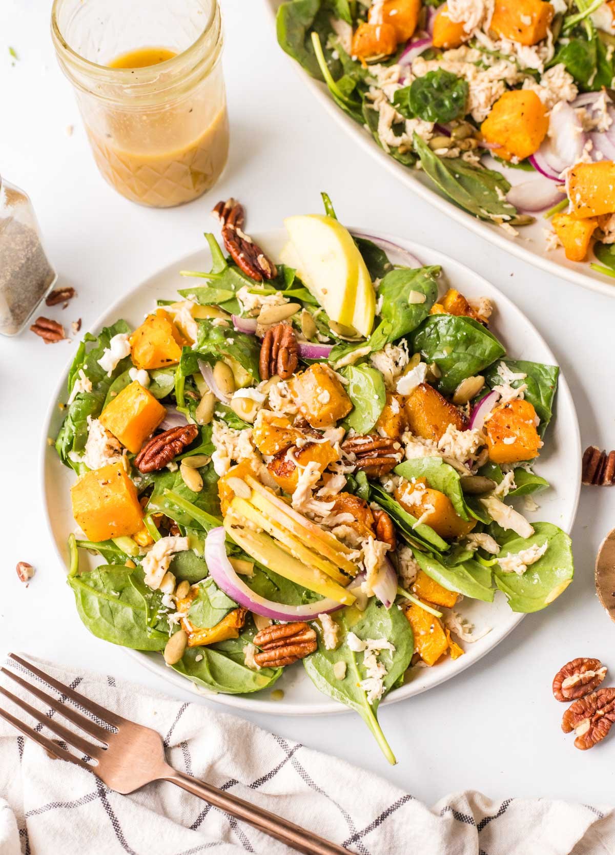 A plate of spinach salad with roasted butternut squash, apple slices, pecans, red onion, shredded cheese, and a jar of dressing on the side.