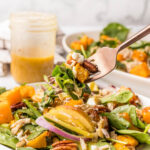 A fork lifts a bite of salad with spinach, roasted butternut squash, pecans, red onions, and dressing. A jar of dressing and a plate of salad are in the background.